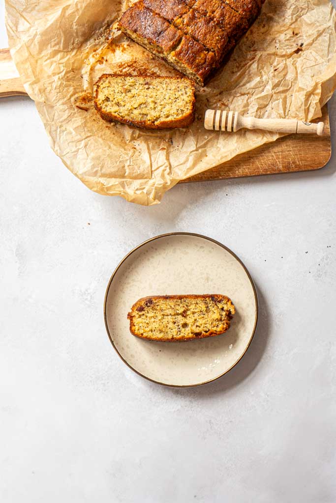 a slice of banana bread on a plate, above it there is a wooden tray with few slices of banana bread straight from the oven